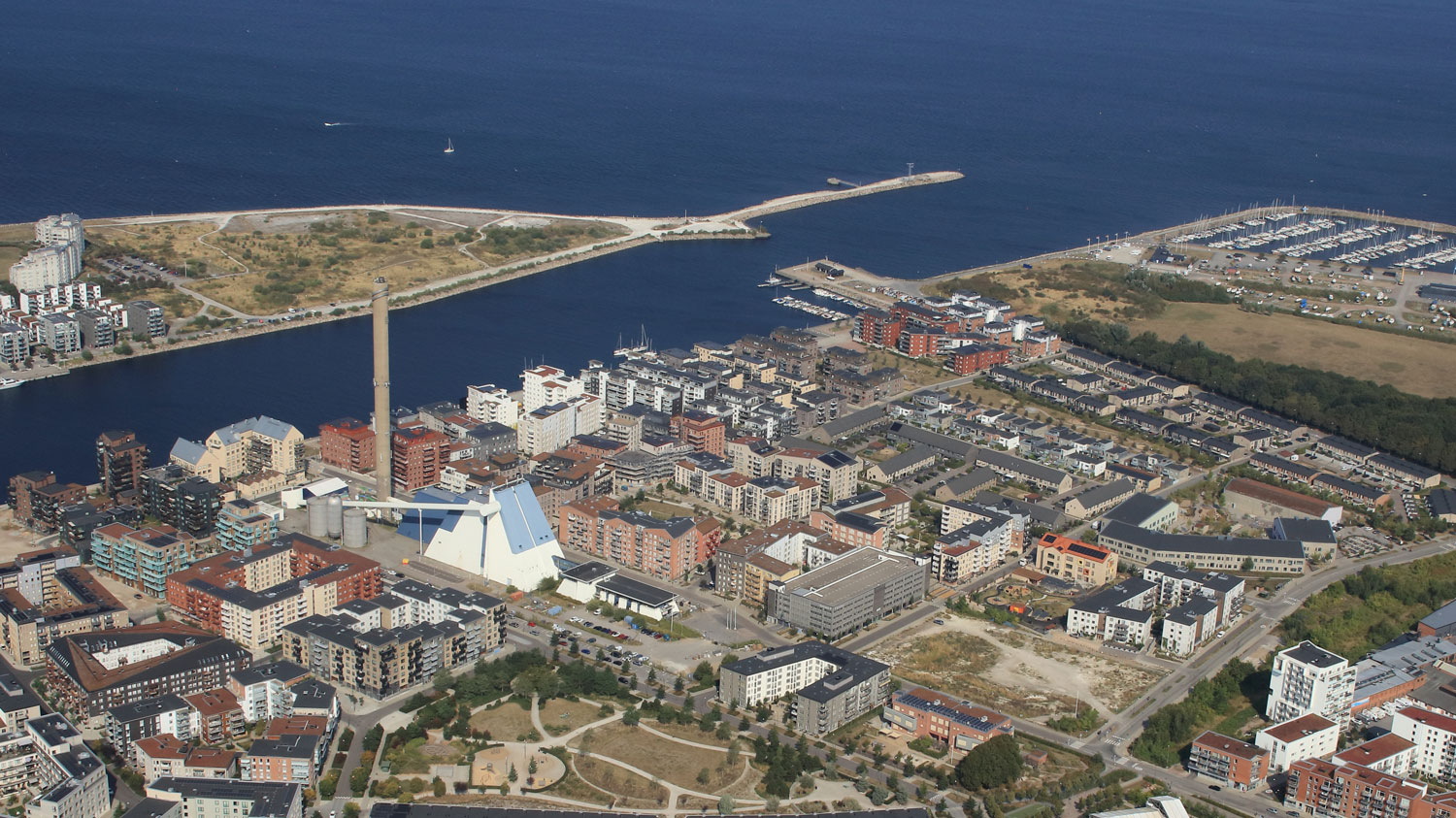 Flygfoto över en urban kustmiljö med bostadshus och parker vid vattnet. Bilden visar en tät stadsbebyggelse med färgglada byggnader, gångvägar, grönområden och en marina i bakgrunden. 