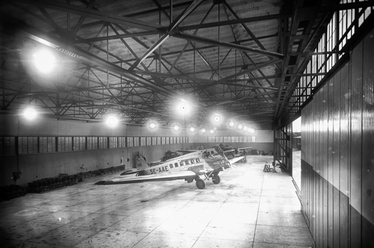 Äldre fotografi på en stor flygplanshangar. I mitten av den stora lokalen står ett vitt propellerflygplan. 