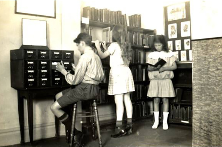 Gammalt svartvitt foto på tre barn i bibliotek.