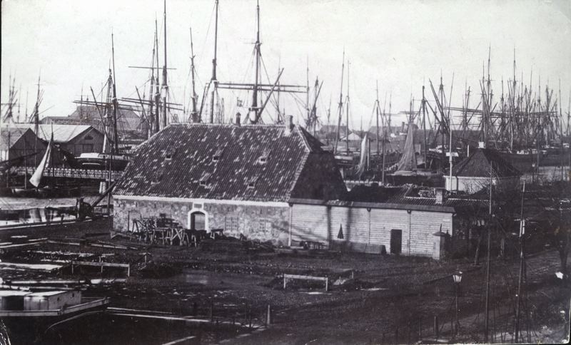 Svartvitt foto av hamnområdet med segelbåtar och byggnader ca 1865-70.