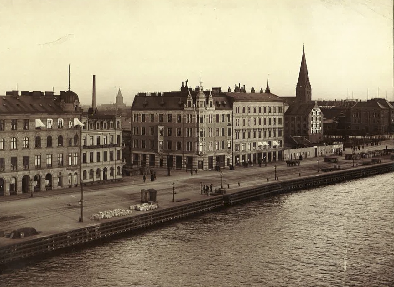 Östra Kajgatan fick sitt namn 1880 och blev Skeppsbron 1911. Kajen var tidigt anläggningsplats för båtar till och från Köpenhamn, såsom hjulångarna Gefion och Gylfe. Husraden byggdes i mitten av 1890-talet. I bildens högra del inhägnas den så kallade Beijerska tomten av ett plank till höger om det är ännu inte posthuset uppfört.