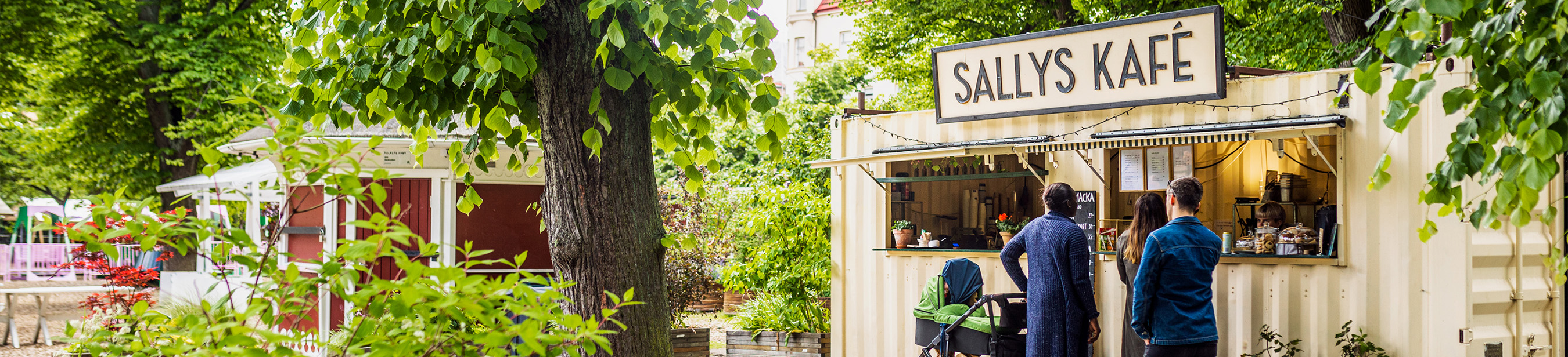 Gröna träd och buskar runt en gul café-byggnad med skylten Sallys Kafé. 