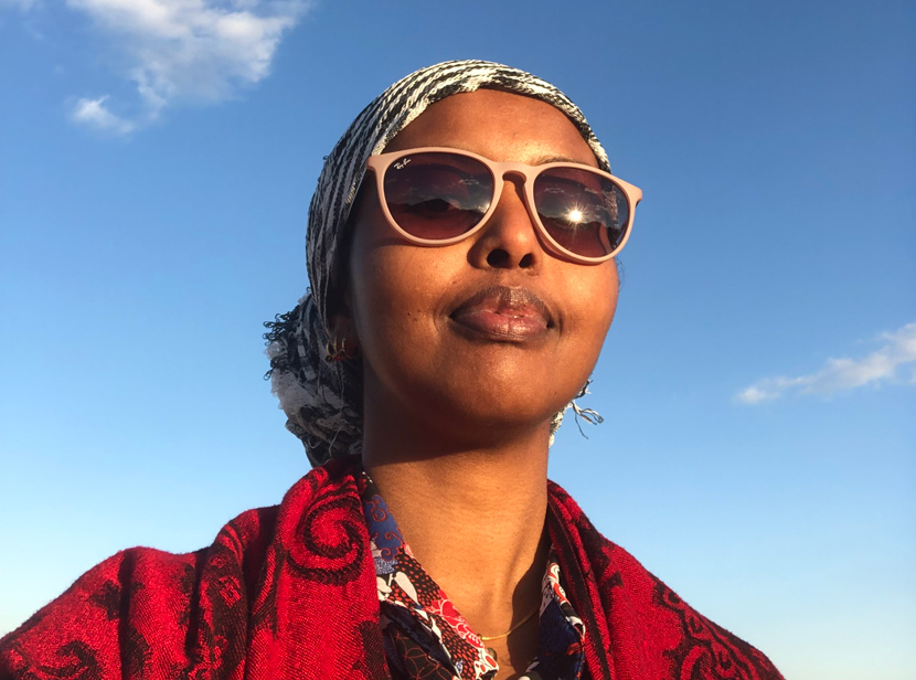 Ayaan profil avtecknar sig mot en blå himmel. Hon har solglasögon och en röd sjal.