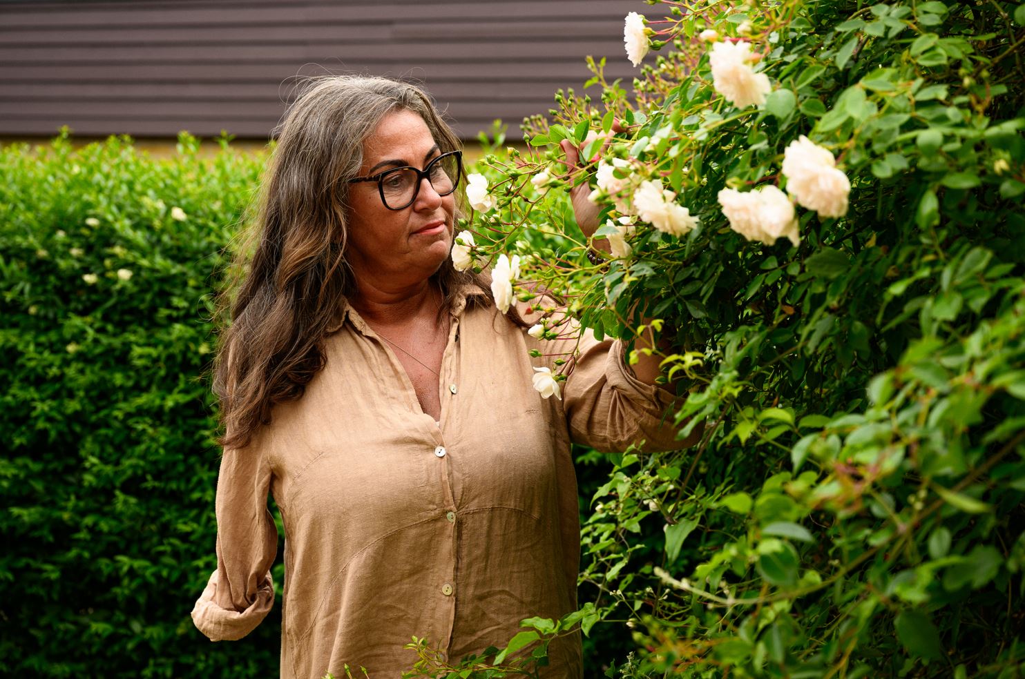 Kvinna med långt mörkt, lite vågigt, hår står vid en rosenbuske med ljusa rosor och håller i en gren med blommor 
