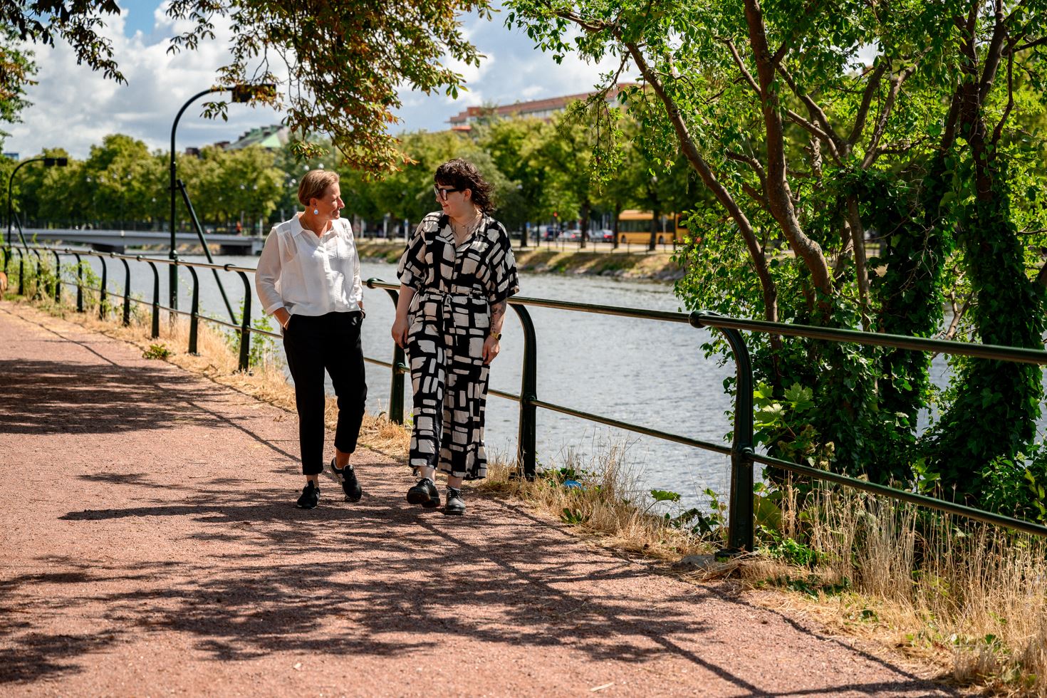 Två kvinnor som ler mot varandra promenerar längs kanalen i Malmö, solen lyser och träden har gröna blad