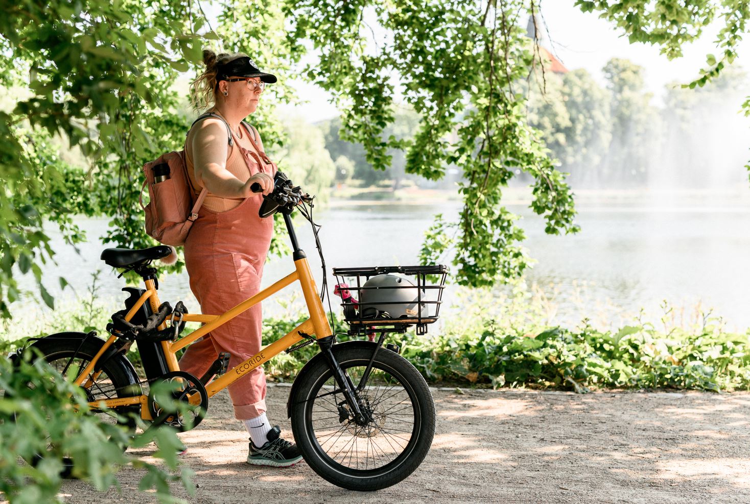 Kvinna med rosa hängselbyxor, rosa ryggsäck och svart skärmkeps promenerar vid en damm och leder en orange-gul cykel. Grönskande träd och växter syns i för- och bakgrunden.
