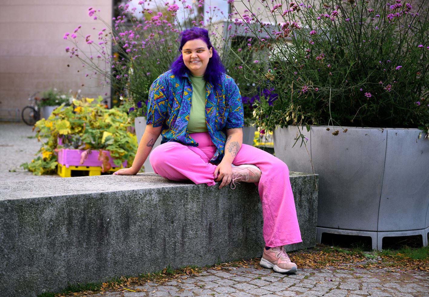 Leende person med lila hår, mönstrad färgglad skjorta, grön t-shirt, rosa byxor och skor, sitter på en stenbänk utomhus, med stora krukor med lila blommor i bakgrunden.