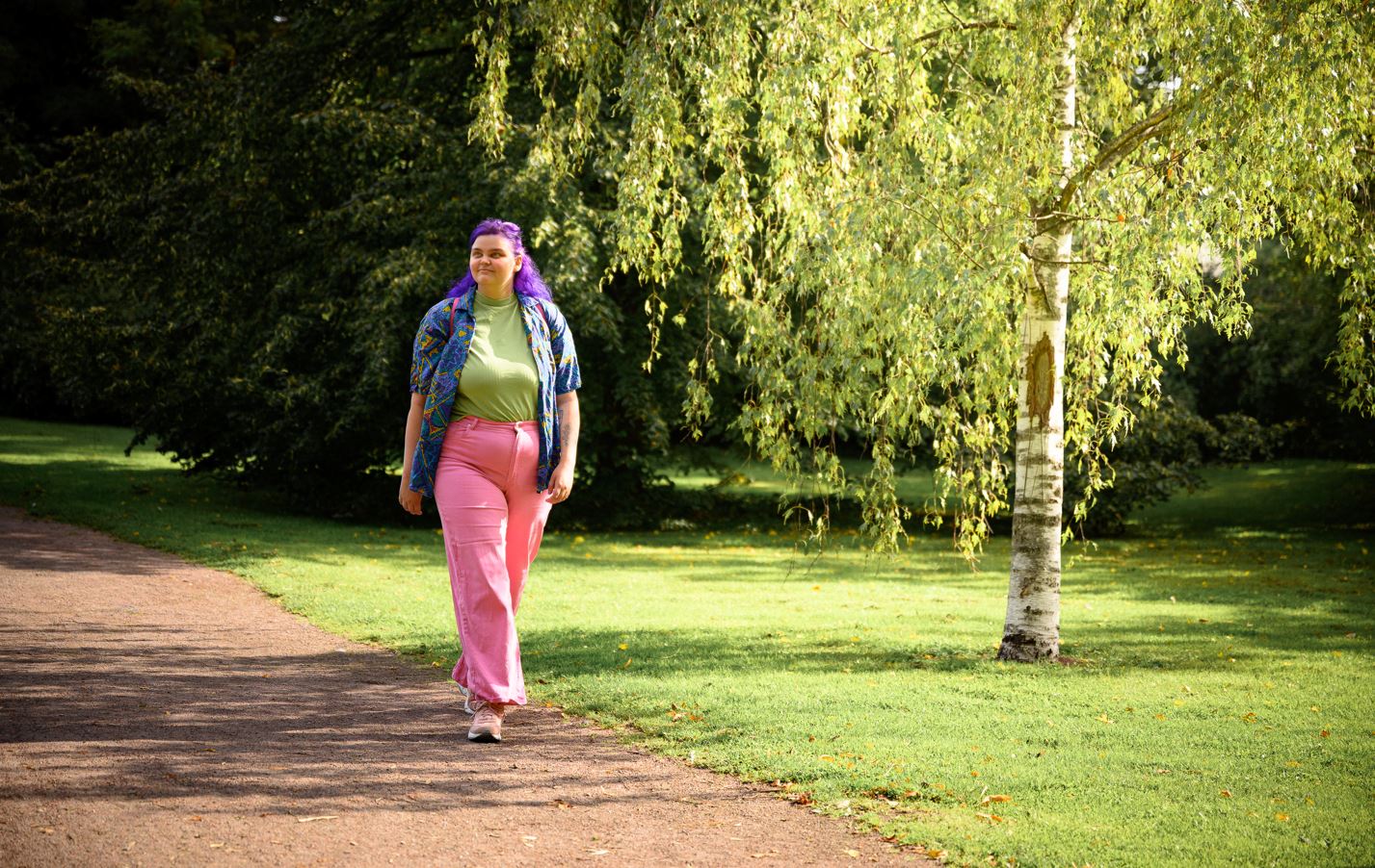 Person med lila hår, mönstrad färgglad skjorta, grön t-shirt, rosa byxor och skor, går i en park och blickar uppåt, i bakgrunden växer lövträd