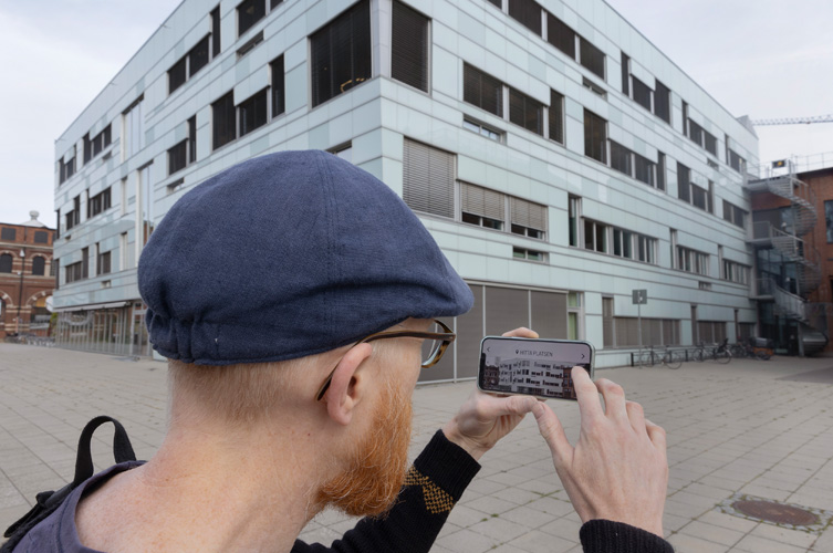 Fotografi taget bakifrån på en man i keps. I handen håller han en mobiltelefon som visar en byggnad på skärmen. Samma byggnad har han också framför sig. 