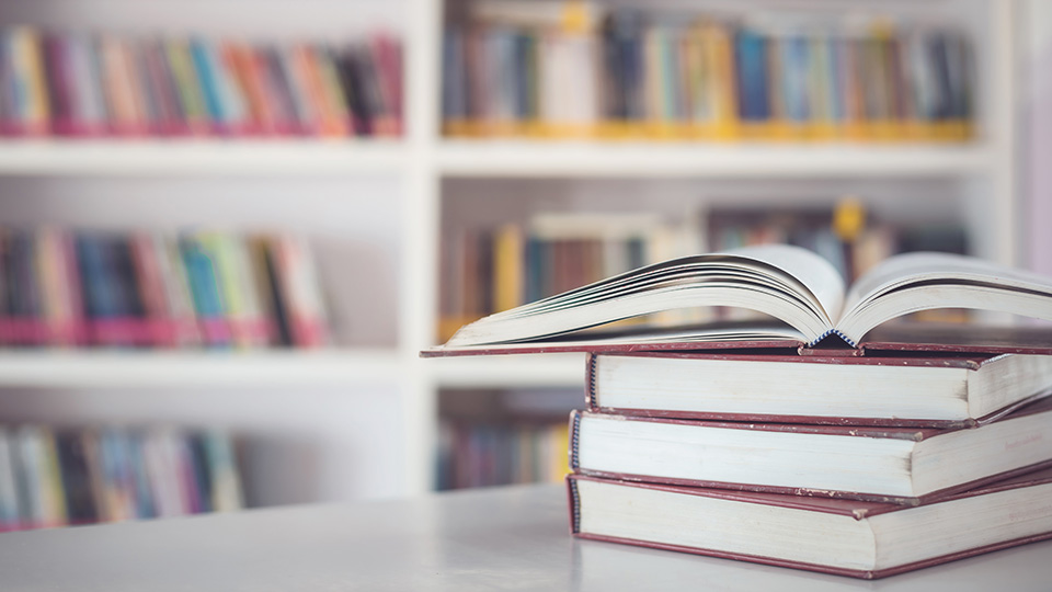 En trave böcker ligger på ett bord. Den översta boken är uppslagen. I bakgrunden syns bokhyllor fyllda med böcker.