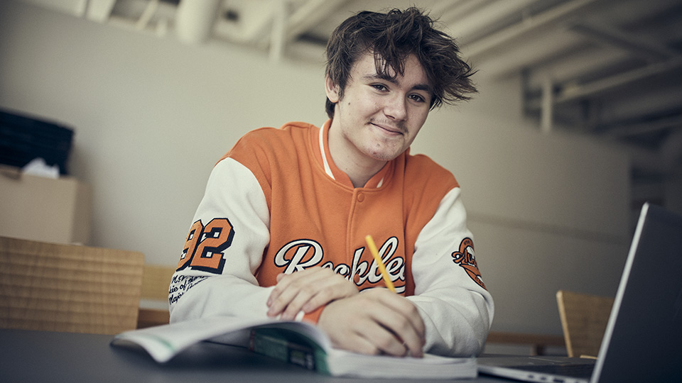 En ung kille med orange tröja sitter i en skolsal. Han håller en penna i handen och skriver i en bok.