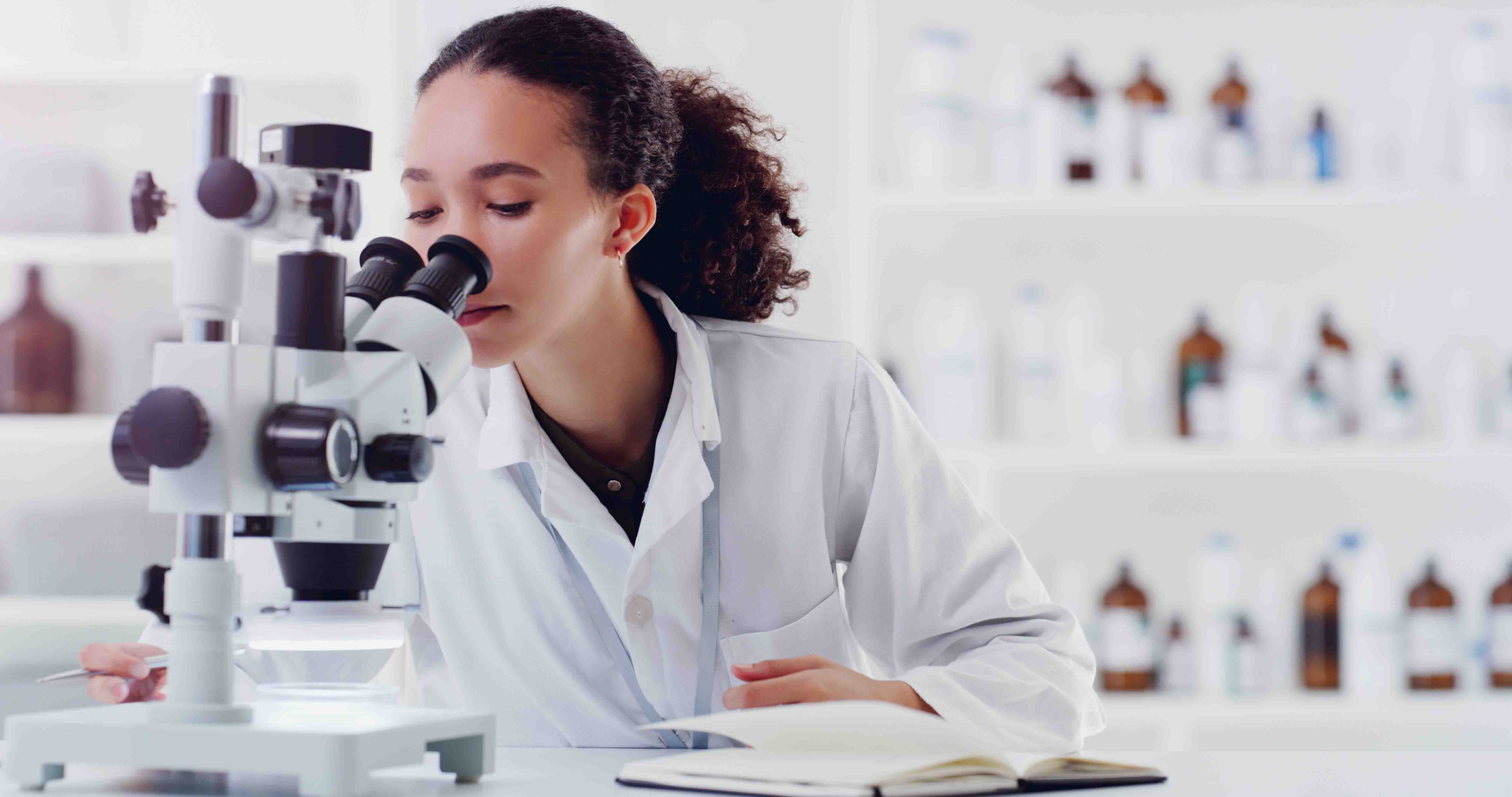 En mörkhårig flicka tittar i ett mikroskop. Hon har vit rock på sig och är i ett laboratorium.