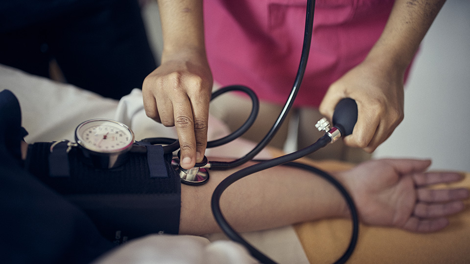 Närbild på en person som mäter blodtrycket på en annan person. Det är endast armar och händer som syns på personerna. 