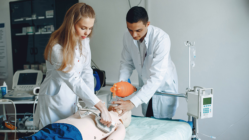 Två elever i sjukvårdskläder övar hjärtstart på sjukvårdsdocka.