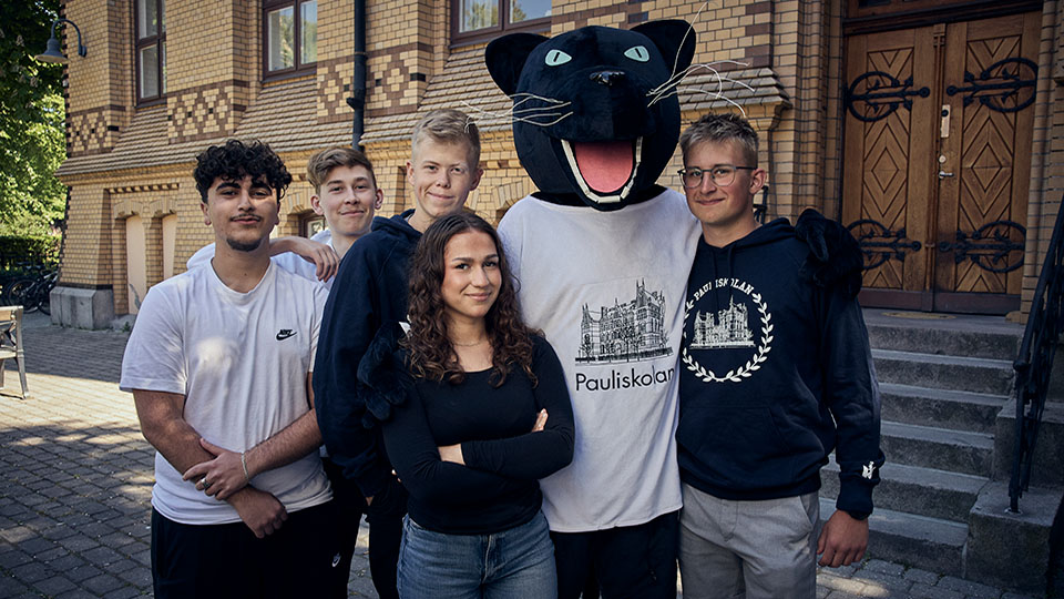 Gruppbild på fem elever och en elev utklädd till skolmaskot utanför skolans entré.