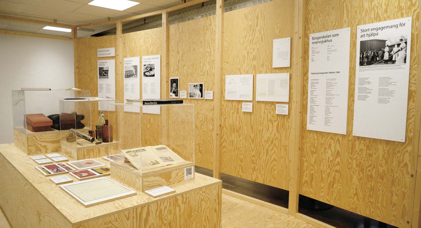 En utställningslokal där vita informationstavlor med text och svartvita bilder hänger på väggar av ljust trä.