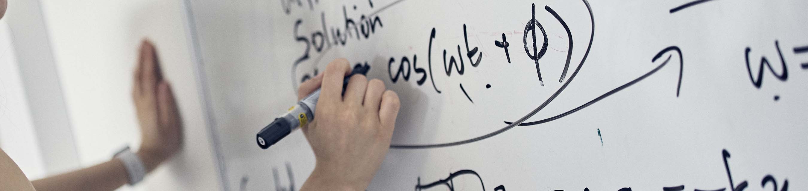 Närbild på en hand som håller på attt skriva en matematisk formel på en vittavla med en svart penna.