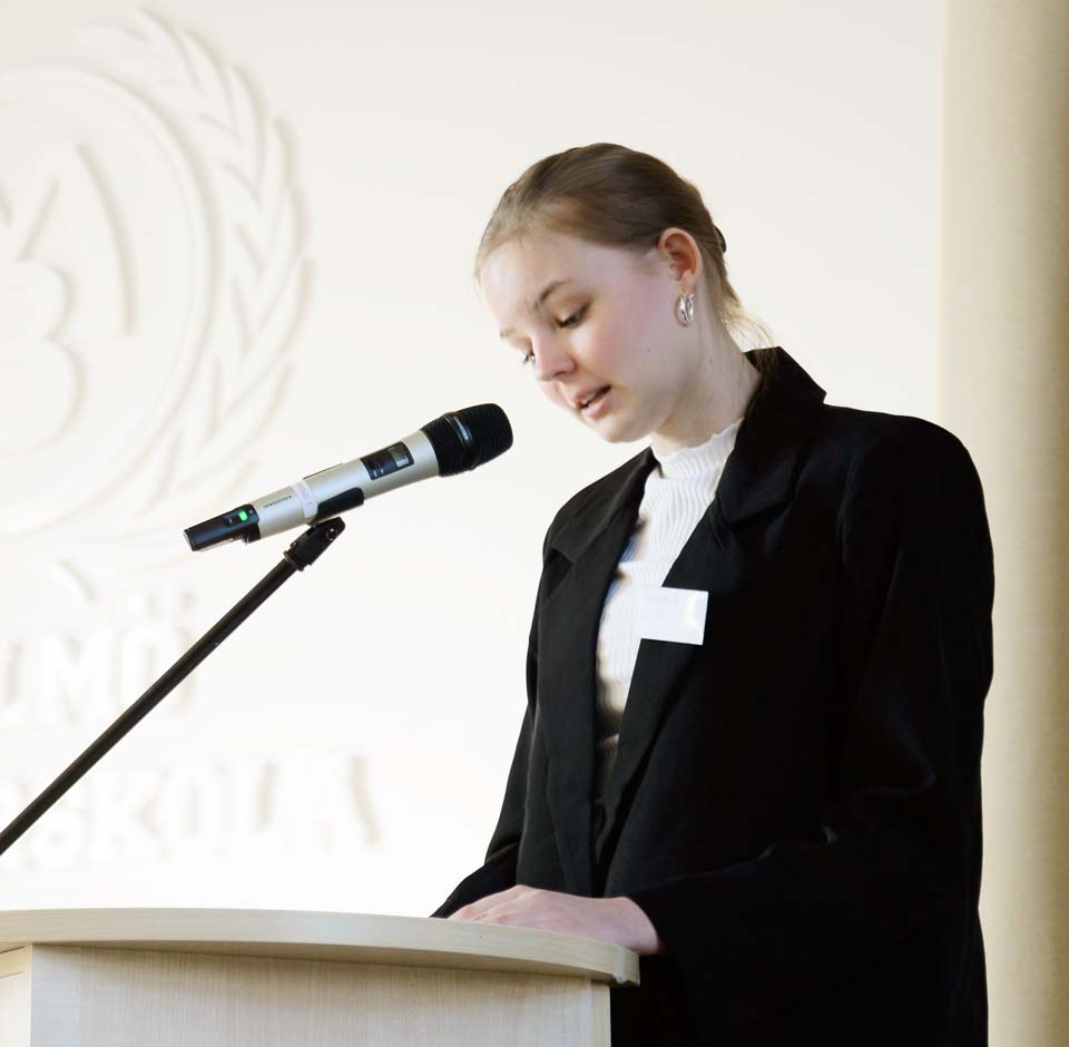 En elev iklädd vit blus och svart kavaj står vid en mikrofon och talar i en ljus inomhusmiljö.