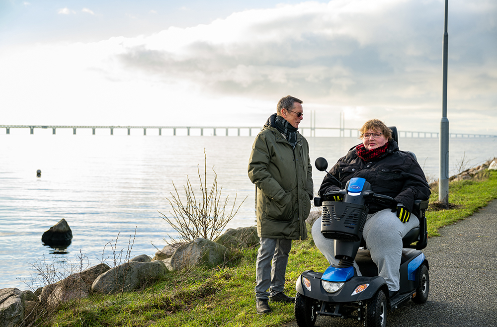 En kvinna i rullstol bredvid en man som står upp och tittar på henne. De har på sig tjocka jackor, i bakgrunden syns havet och Öresundsbron.