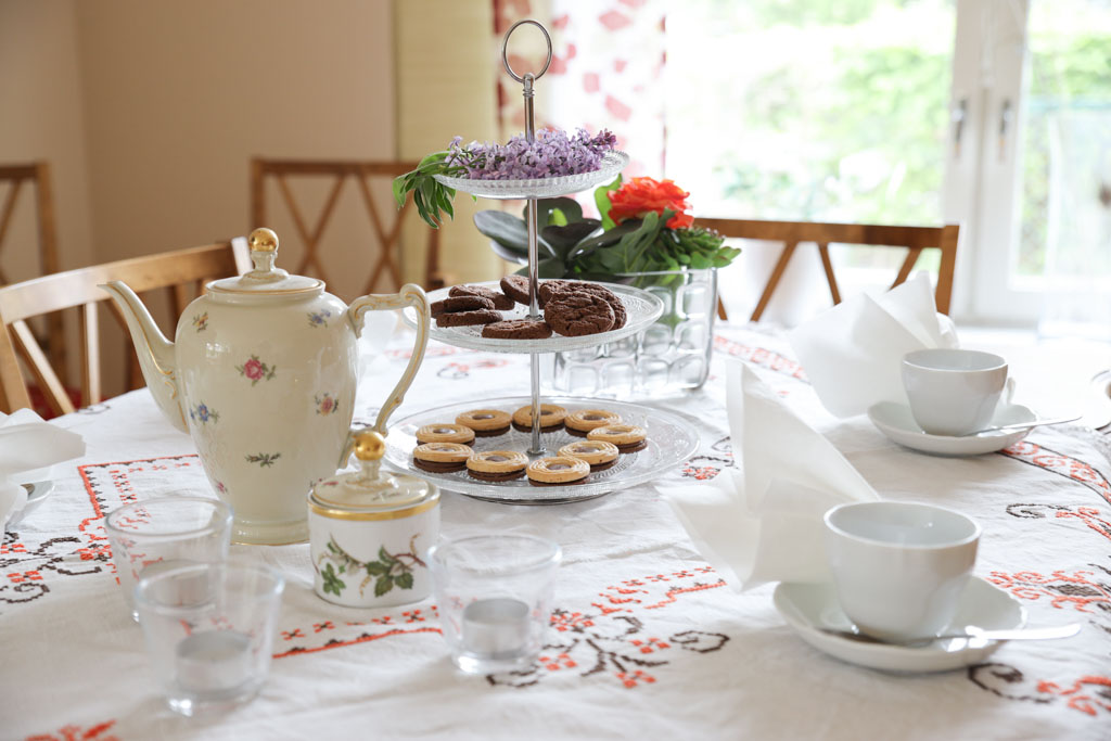 Ett uppdukat bord med linneduk, kakor och finporslin bjuder in till fika.