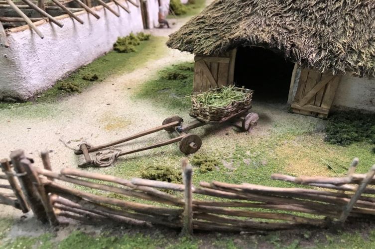 Miniatyrmodell av släpvagn från järnålder. Samt omgivande byggnader, tomt och staket.