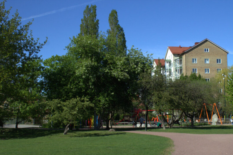 Park med höga träd omringad av flerfamiljshus.