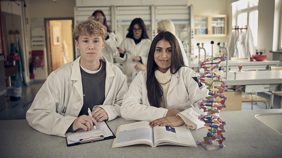 Två elever i labbsalar tittar rakt in i kameran och ler. I bakgrunden syns elever som laborerar i laboratoriesal på skolan.