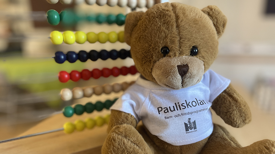 Närbild på en teddybjörn. I bakgrunden syns en kulram. Teddybjörnen bär en vi tröja med texten Pauliskolan.