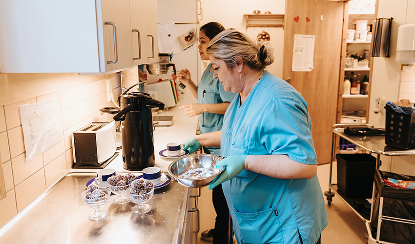 Dijana och Puangpaka står i köket i sin blåa jobbklädsel.