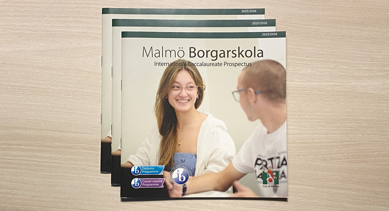 Three copies of a printed booklet lie in a pile on a light-coloured surface. On the cover, a photograph shows two smiling students seated next to one another in a classroom setting.