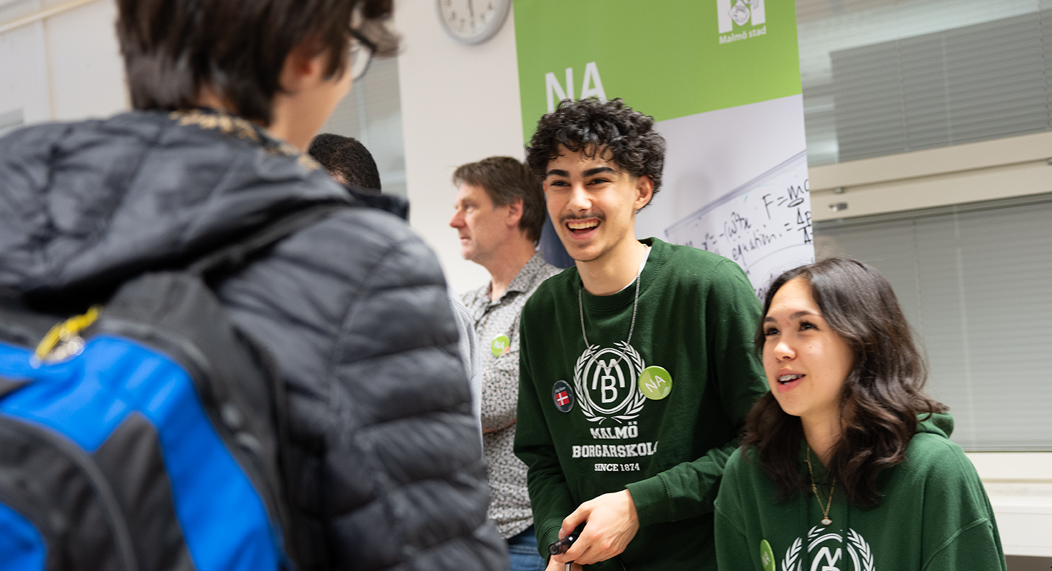 Två elever med gröna tröjor med Borgarskolan-motiv pratar med en besökare under Öppet hus.