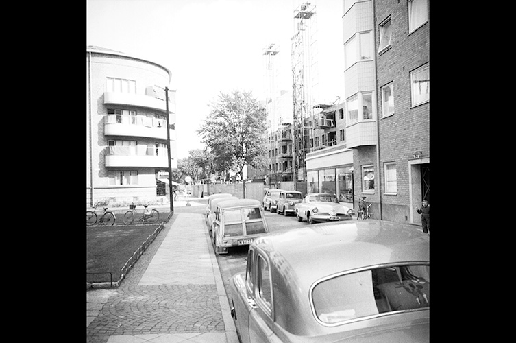 Svartvitt foto av gata med hus längs sidorna och bilar parkerade längs trottoaren.