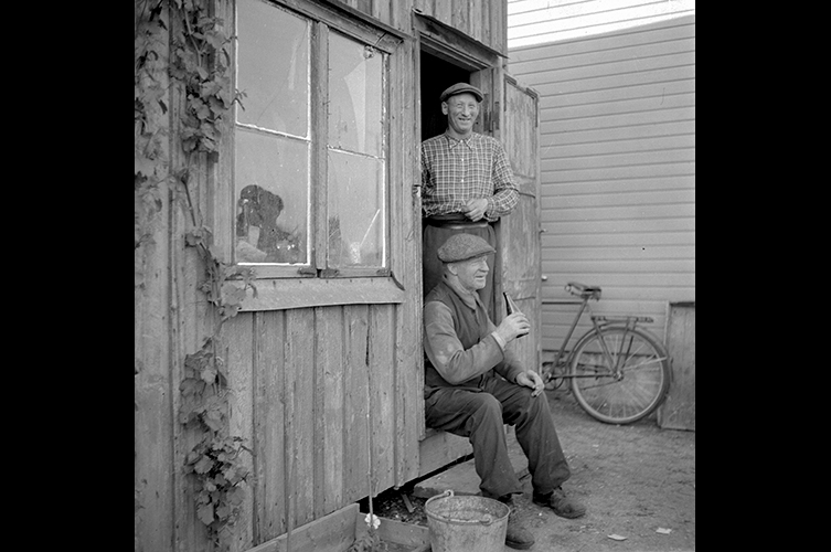 Äldre fotografi på två män. Den ena mannen står upp i dörröppningen till en hodda, och den andra sitter ner i dörröppningen och håller en glasflaska i sin hand.