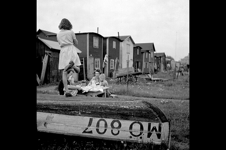 En flicka står på en uppochnervänd eka, bakom henne sitter personer vid ett bord och i bakgrunden små trähus.