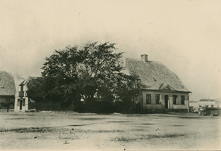Äldre fotografi på Hamnmästarbostället. Framför huset står ett stort träd och i bakgrunden syns hamnen. 