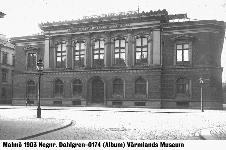Äldre fotografi på gamla Riksbanken, en stor byggnad i två våningar. Ovanvåningen har stora fönster. Högst upp ovanför fönstren står det "Riksbankens Afdelningskontor"