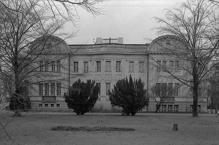 Äldre fotografi på Fridhemsborg, en pampig byggnad med kolonner. Framför byggnaden ligger en stor gräsmatta, med buskar och träd. 
