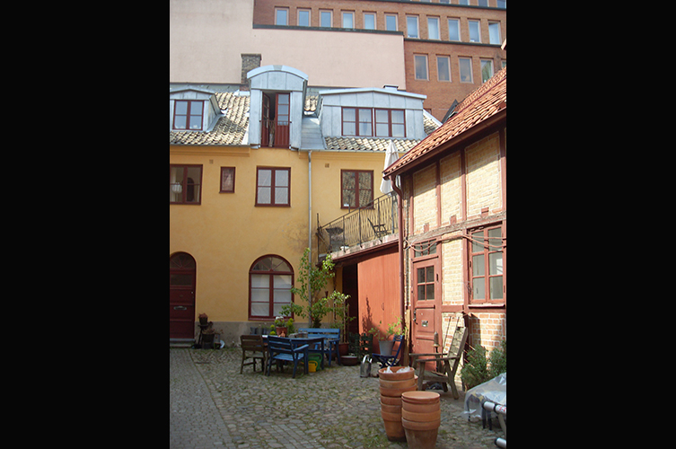 Fotografi på en innergård med kullersten, framför ett gult hus står utemöbler.