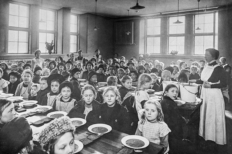Äldre fotografi på barn vid tre långbord. Framför sig har de tallrikar med en gryta eller soppa. Vid det ena bordets kant står en kvinna i vitt förkläde. 