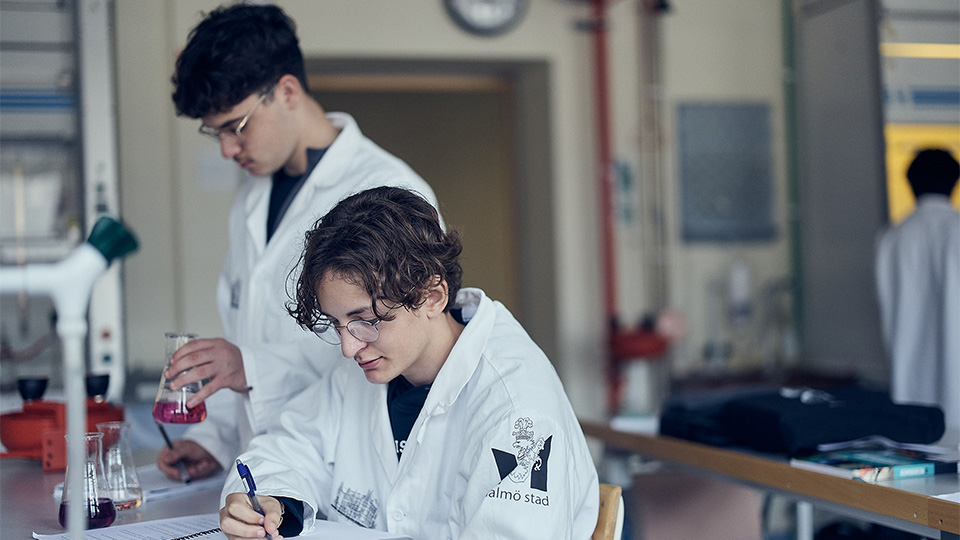 Två elever i vita labbrockar gör laboration i skolans labbsal.