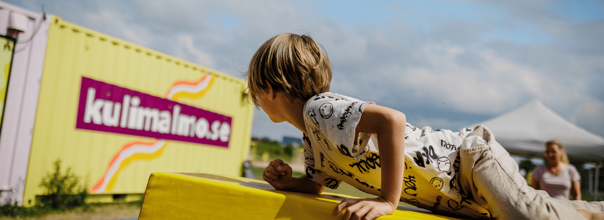 Ett barn klättrar över ett gymnastikredskap. I bakgrunden syns en gul container med texten "kulimalmo.se".