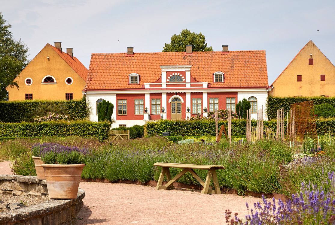 Rabatter med blommande växter och en bänk framför en historisk byggnad som syns i bakgrunden.
