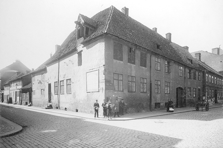 Äldre korsvirkeshus i en korsning, två våningar hög och med flera skorstenar. 