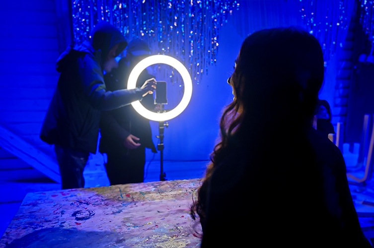 Två personer står vid ett ringljus för filmning, och en person filmas. Starkt ljussatt miljö i blått. 