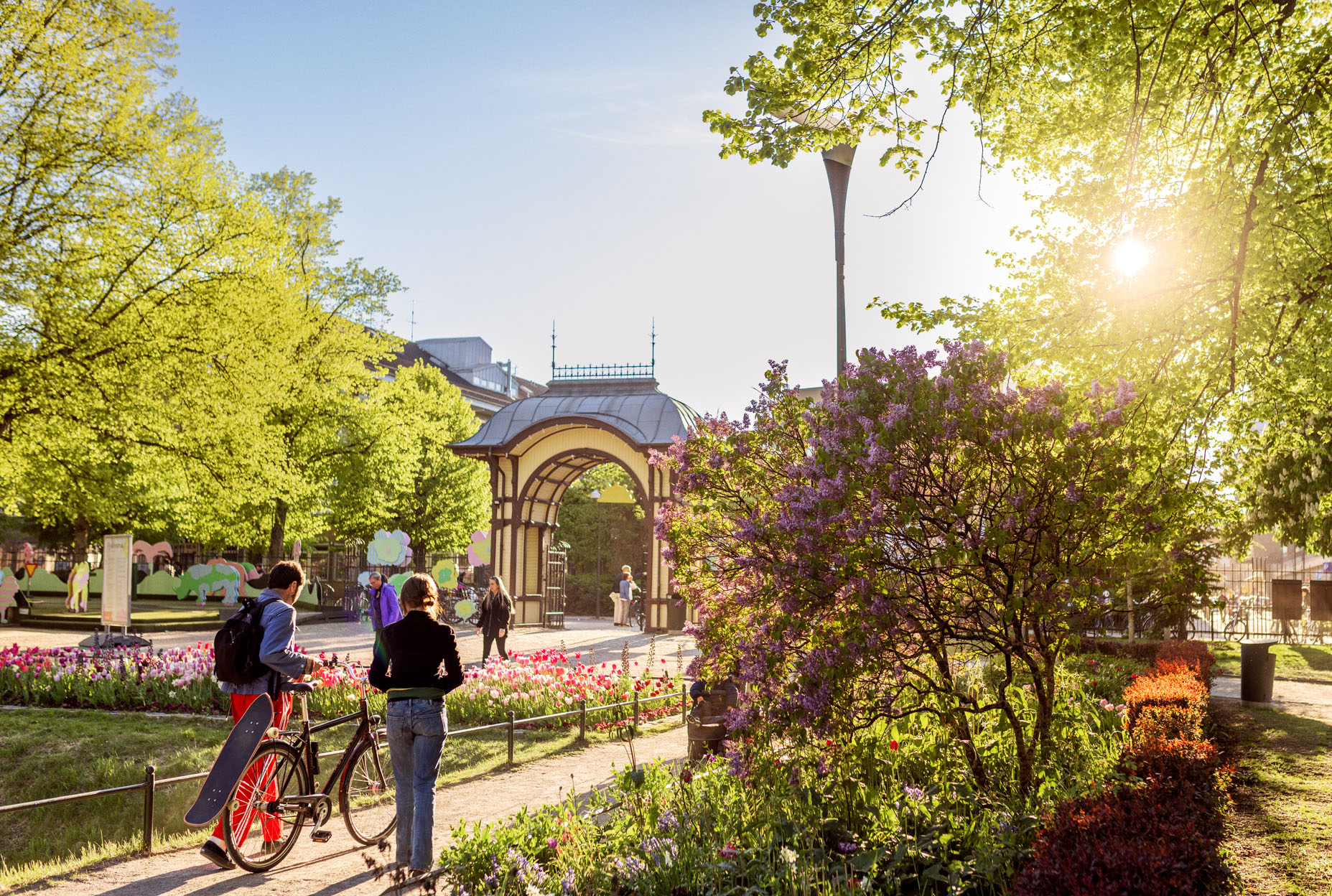 Malmö Folkets Park i kvällssol. Två personer går längs långa dammen, i bakgrunden syns gamla entrén. 