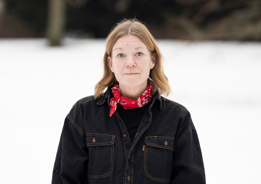 Anna har rött hår, en svart jacka och en röd scarf runt halsen.