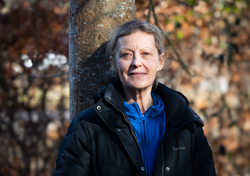 Anna står lutad mot ett träd. Hon har ljust hår, en blå tröja och svart jacka.