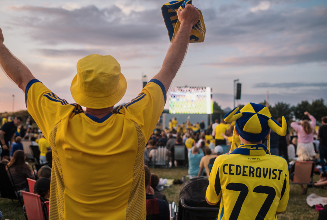 Två personer i landslagströjor för Sveriges fotbollslandslag ser en match på storbildsskärm utomhus tillsammans med flera andra fotbollsfans.