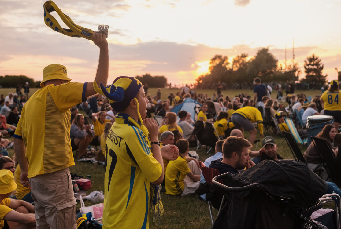 Två svenska fotbollsfans kollar på en match på storbildsskärm bland en mängd andra svenska fans.