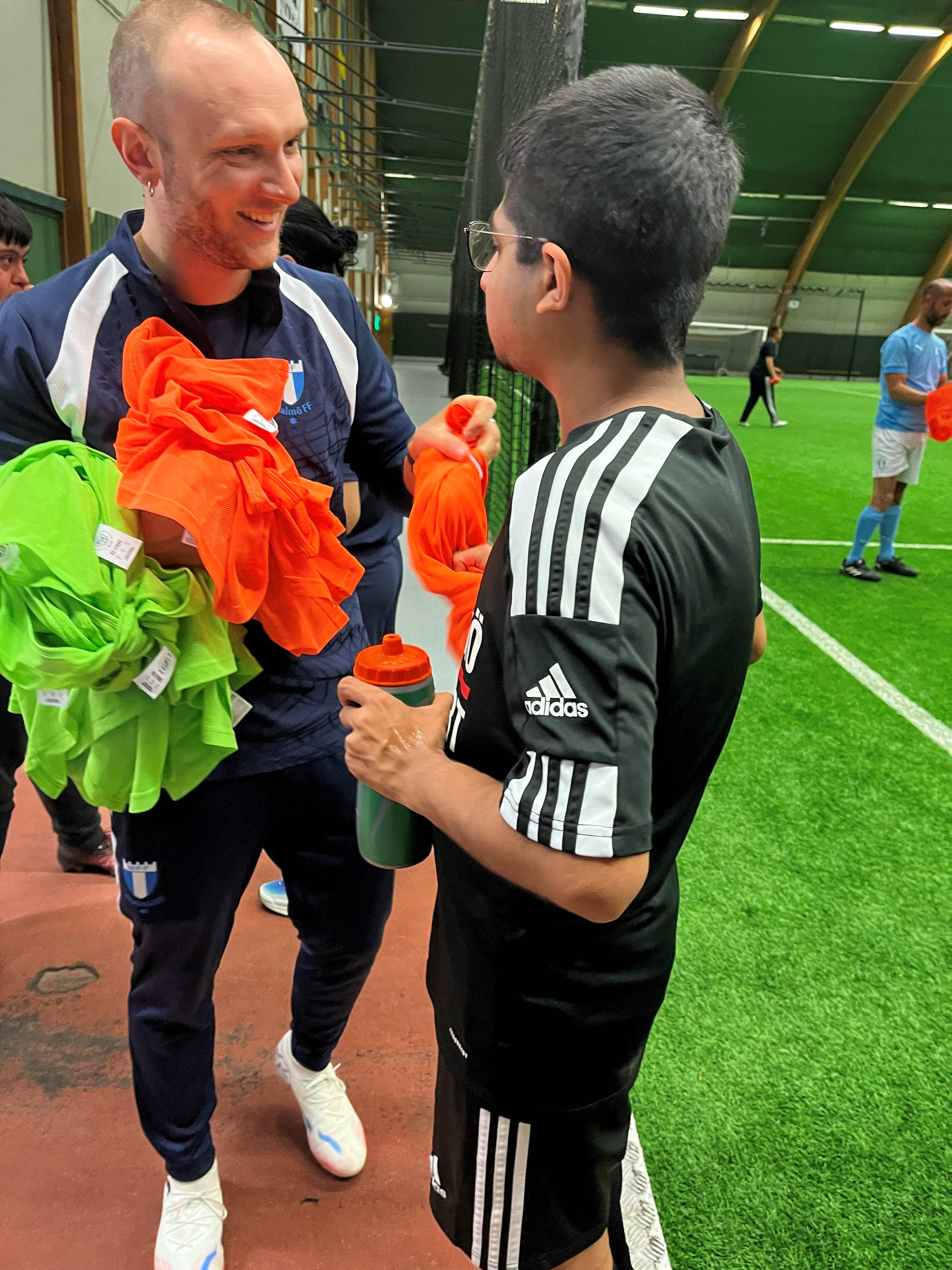 Hamza och Luigi står och pratar med varandra på fotbollsplanen. De är glada.